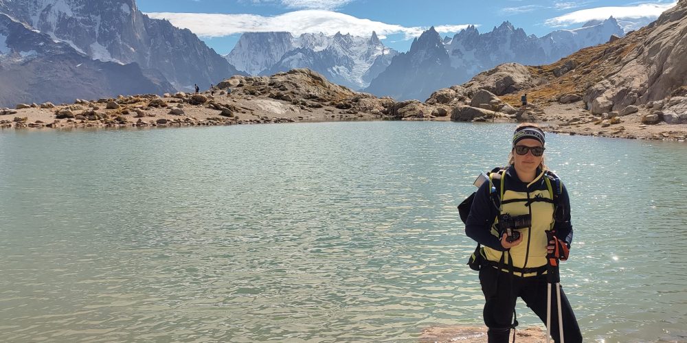 Aurore, en tenue de randonnée et bâtons à la main, au bord du lac Blanc, face aux aiguilles enneigées du massif du Mont-Blanc.
