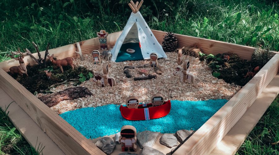 Bac en bois installé dans l'herbe : tipi de tissu, figurines, rivière de riz bleu, canoë rouge, animaux de la forêt, copeaux de bois et mousse.