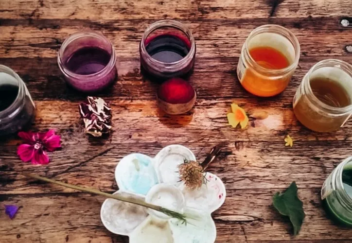 Bocaux en verre remplis de teintures végétales violettes, rouges et jaunes, posés sur une table en bois avec des fleurs et des feuilles.