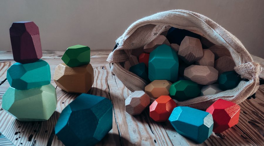 Blocs d'équilibre en bois aux formes irrégulières, colorés en vert, terracotta et bleu, empilés sur une table en bois, le reste dans un sac en toile.