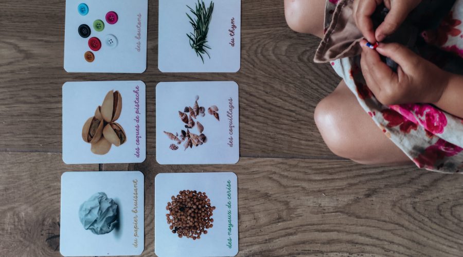 Vue de dessus : un enfant manipule des pochons de tissu colorés à côté de cartes illustrées — un grelot, des boutons, du thym, des coques de pistache, des coquillages, du papier bruissant, des noyaux de cerise.